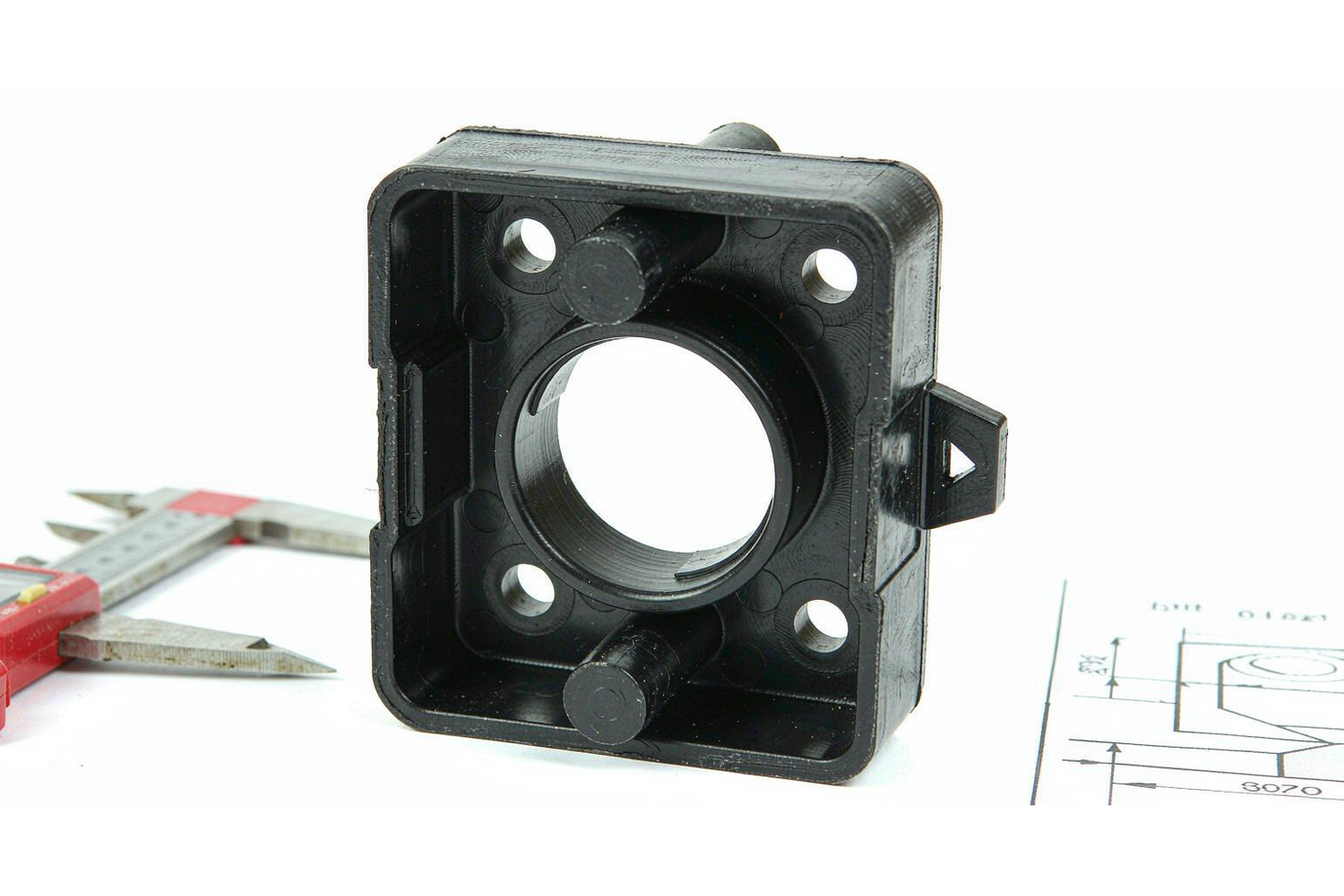 Black ABS molded part measured with a digital caliper beside an engineering drawing during inspection