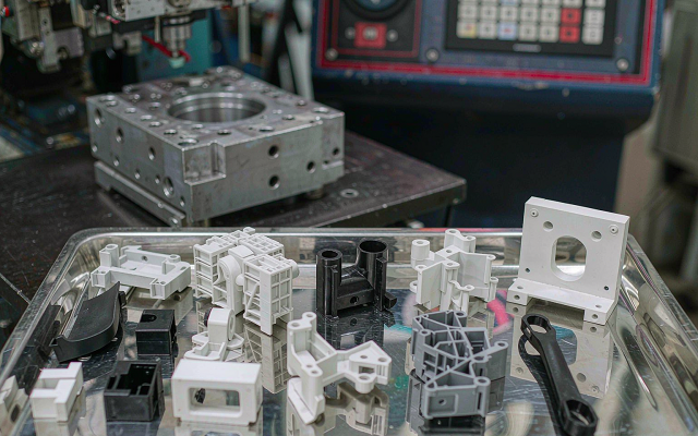 Assortment of injection molded plastic prototype parts in black, white and grey engineering resins laid out on a stainless steel inspection bench with an injection mold in the background