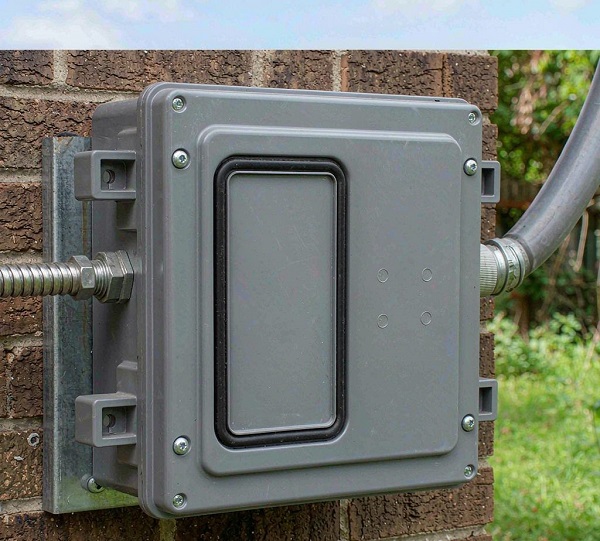 Grey junction box mounted on a brick wall with conduit cables under a blue sky
