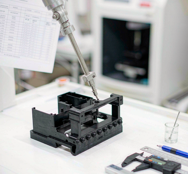 Coordinate measuring machine probing a black injection molded plastic housing part with calipers and inspection documents on a clean bench