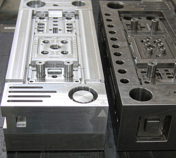 Two injection mold blocks side by side, a bright machined aluminium block and a darker grey pre-hardened steel block, both with visible cavity detail