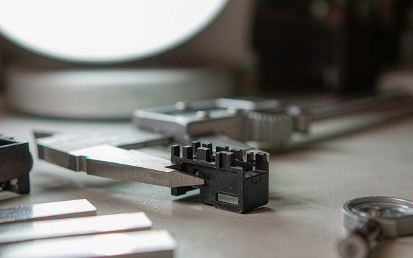 Small black injection molded plastic part being measured with a digital caliper on an inspection bench