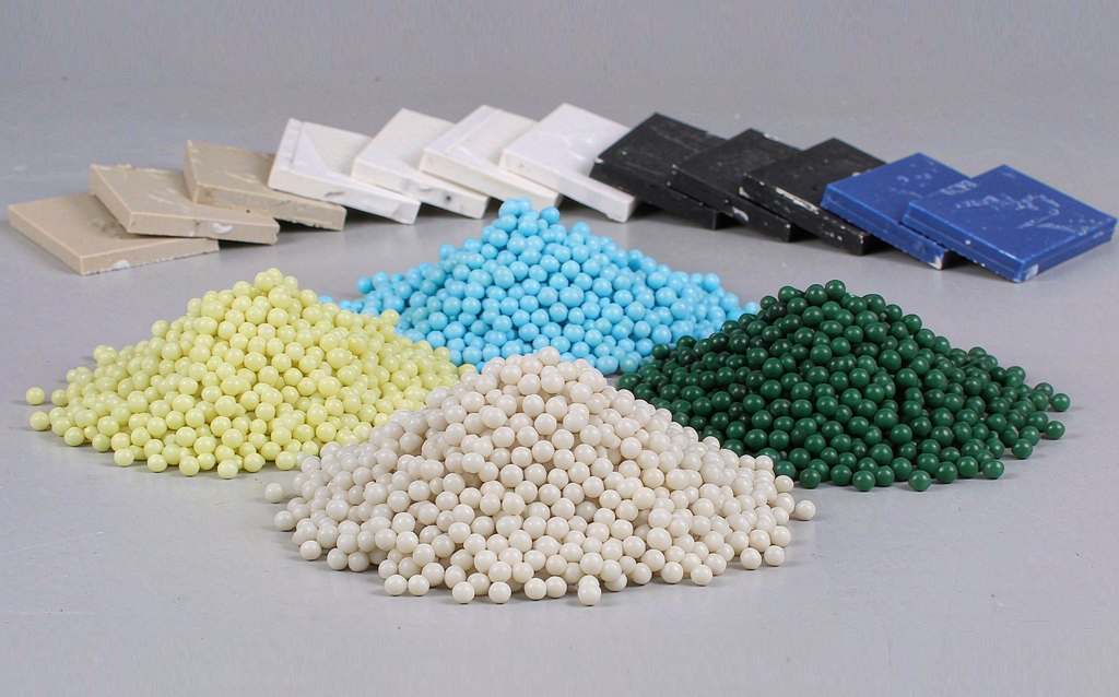 Clear plastic injection molding resin pellets in small piles beside molded plastic test plaques in black, white, natural and blue