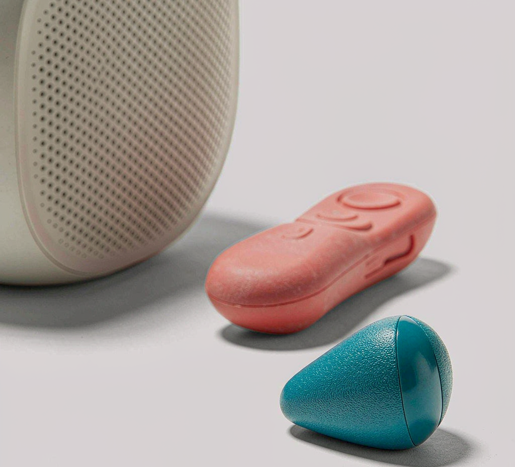 Group of injection-molded consumer electronics housings — off-white smart speaker shell with perforated grille, coral red remote control shell and teal IoT sensor cover — arranged on a light studio surface