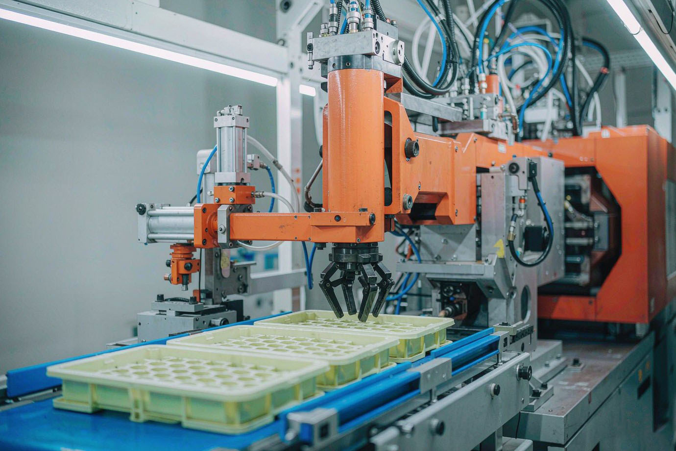 Illustrative — bright injection molding machine producing trays of freshly molded industrial plastic components
