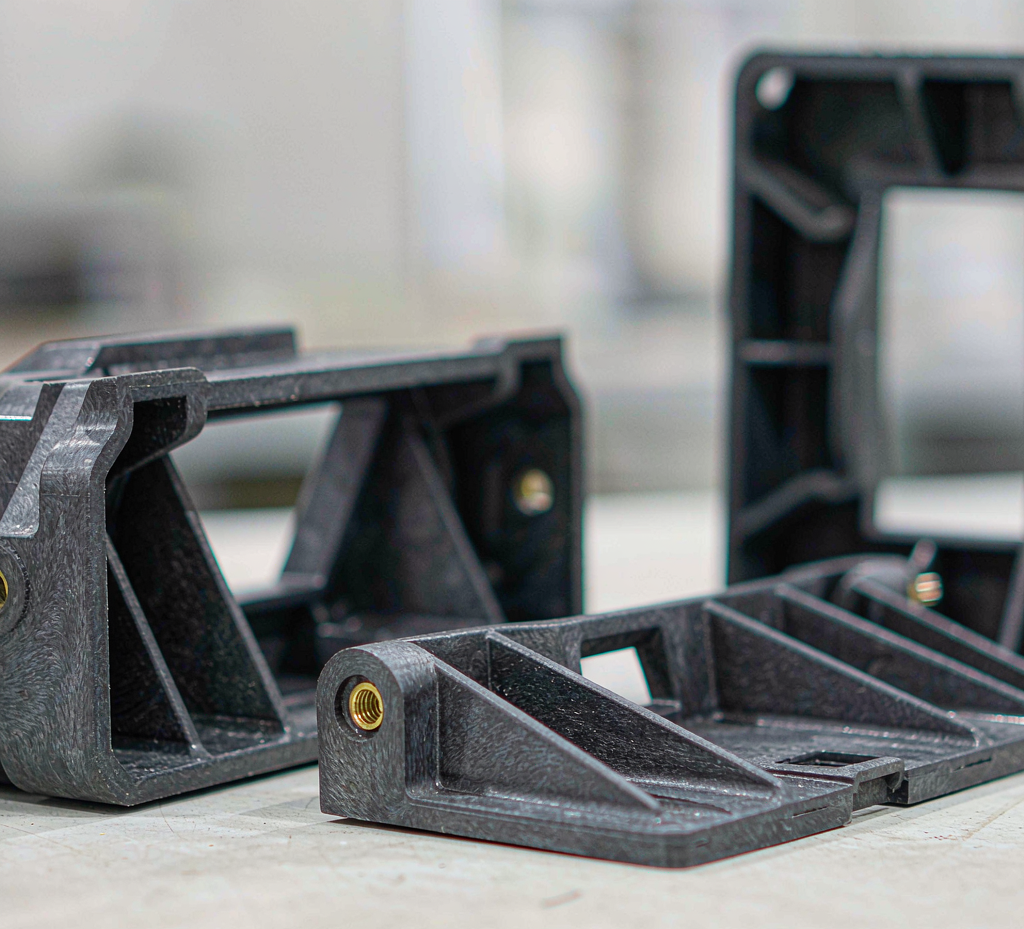 Injection-molded glass-fiber reinforced structural parts — ribbed bracket with threaded insert, mounting base and support frame — on a factory workbench