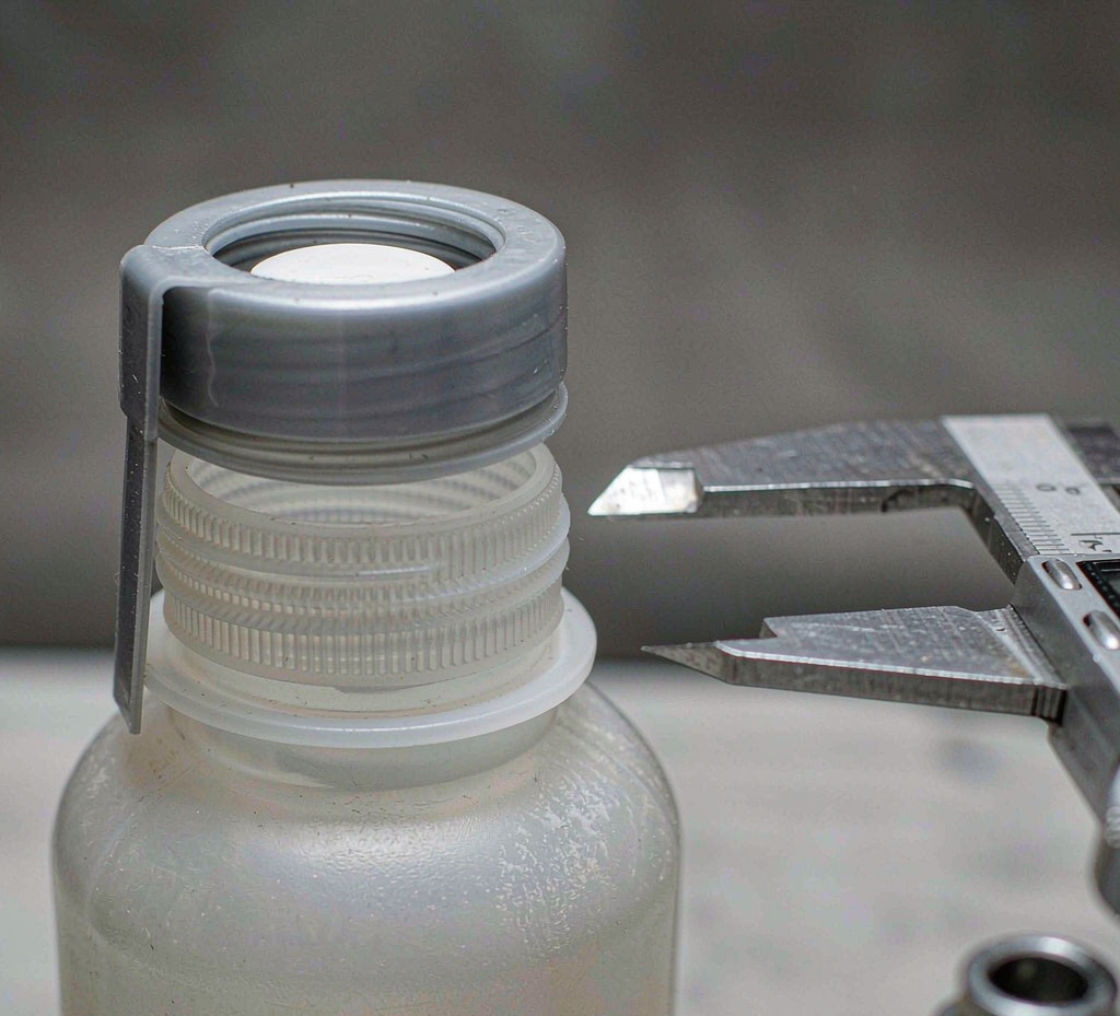 Close-up of a pharmaceutical bottle neck finish with threaded rim, tamper-evident ring on the cap and sealing liner beside calipers