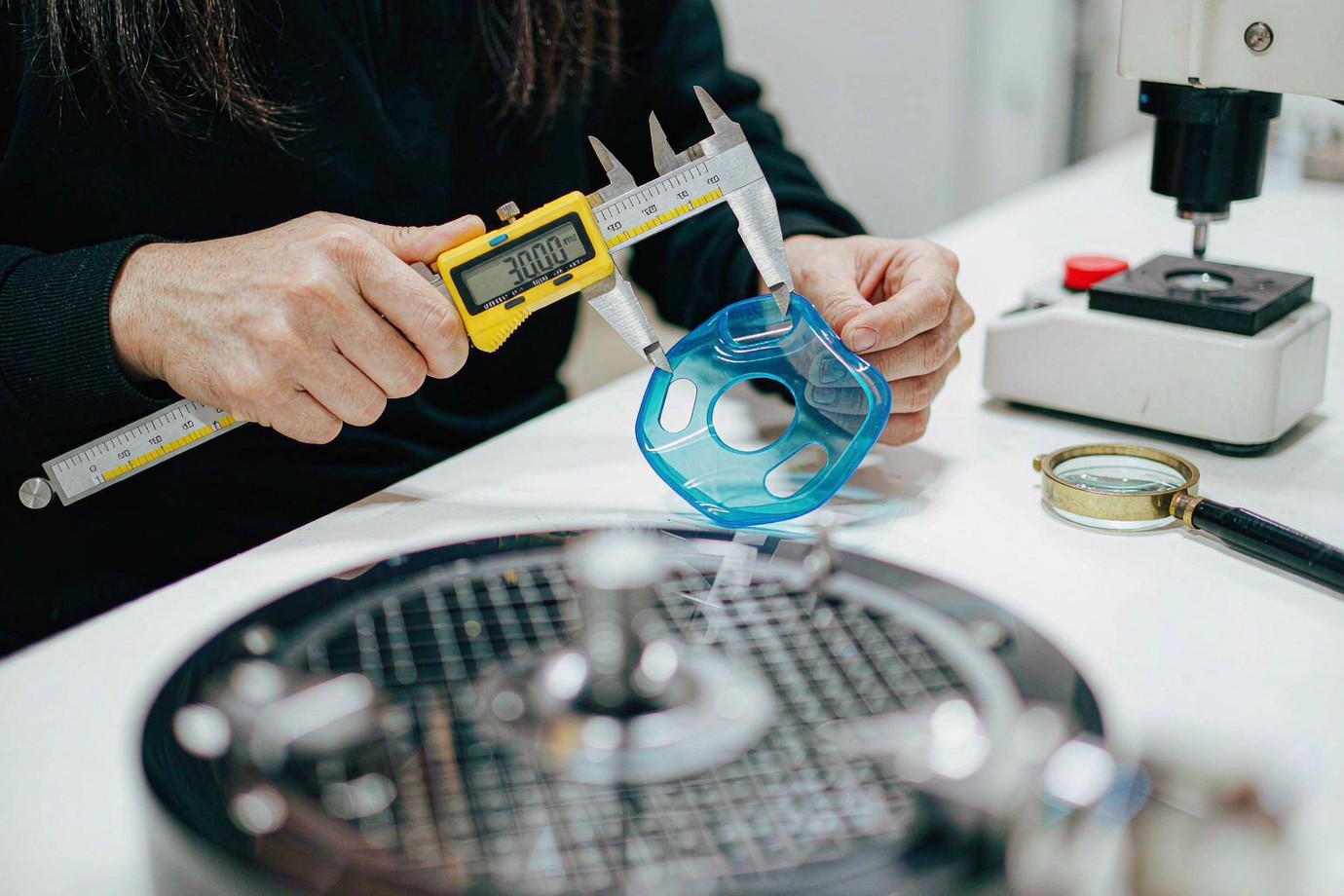 Illustrative — close-up inspection of a translucent blue silicone part with calipers and a hardness tester on a bright bench