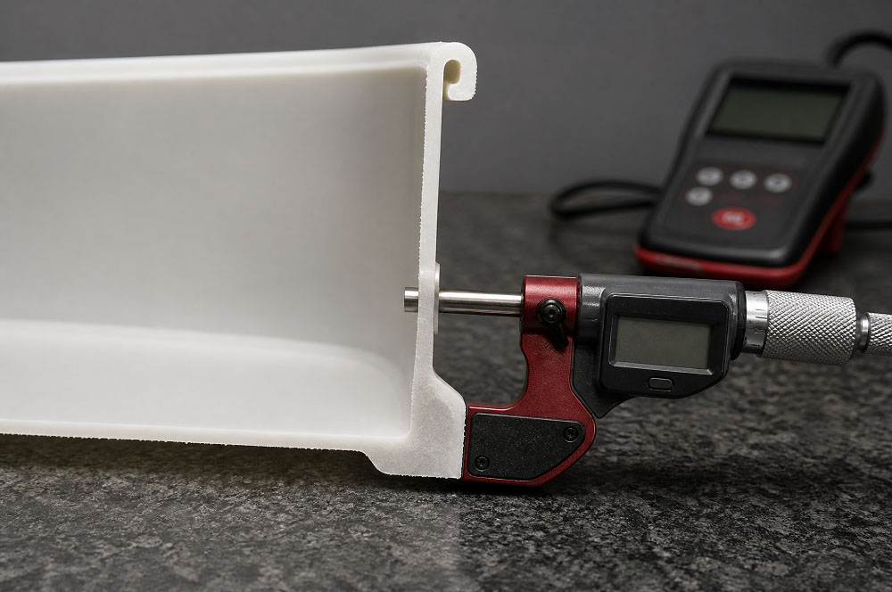 A white polypropylene container cut through its mid-plane and resting on a granite surface plate, the cut edge showing a thin side wall, a much thicker base corner and a rolled rim bead, with a digital micrometer on the wall and an ultrasonic wall-thickness gauge behind it