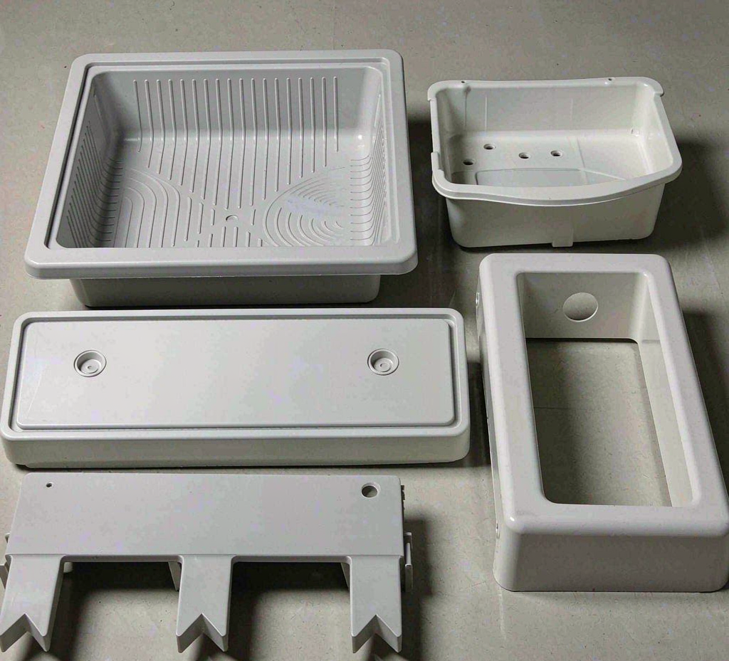 Injection-molded plastic components for major home appliances — crisper drawer, detergent dispenser housing, control panel bezel, handle segment and mounting bracket — on a factory workbench