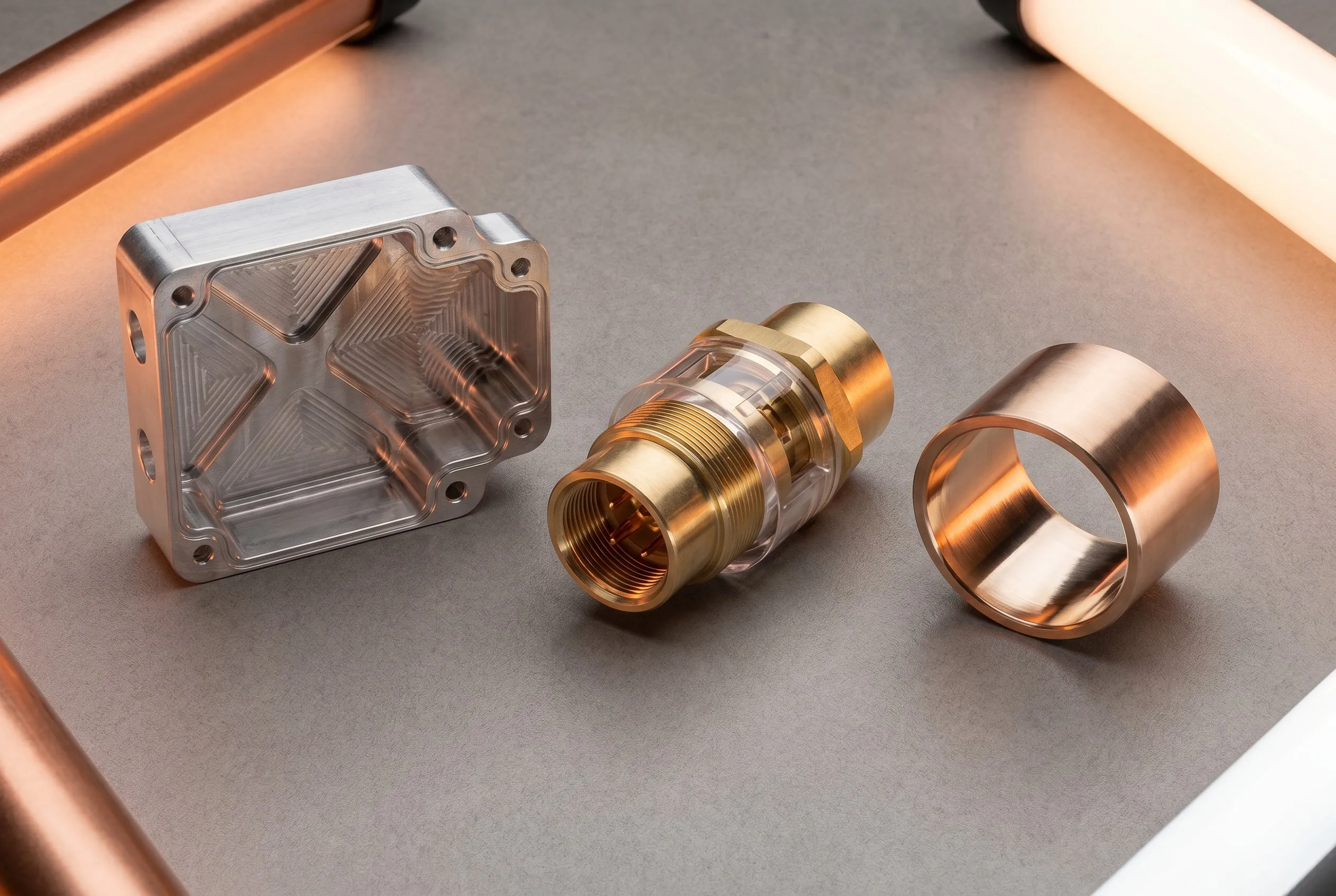 Three CNC machined metal parts: aluminum robotics housing, brass electrical connector, bronze bearing sleeve